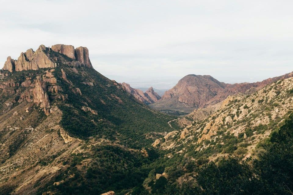 A rugged mountain range with a prominent rocky peak overlooks a lush, green valley under an overcast sky.