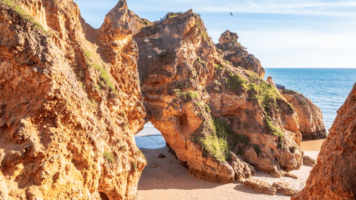 Formazioni rocciose arancioni baciate dal sole creano un arco naturale su una spiaggia sabbiosa, con il placido oceano blu sullo sfondo.