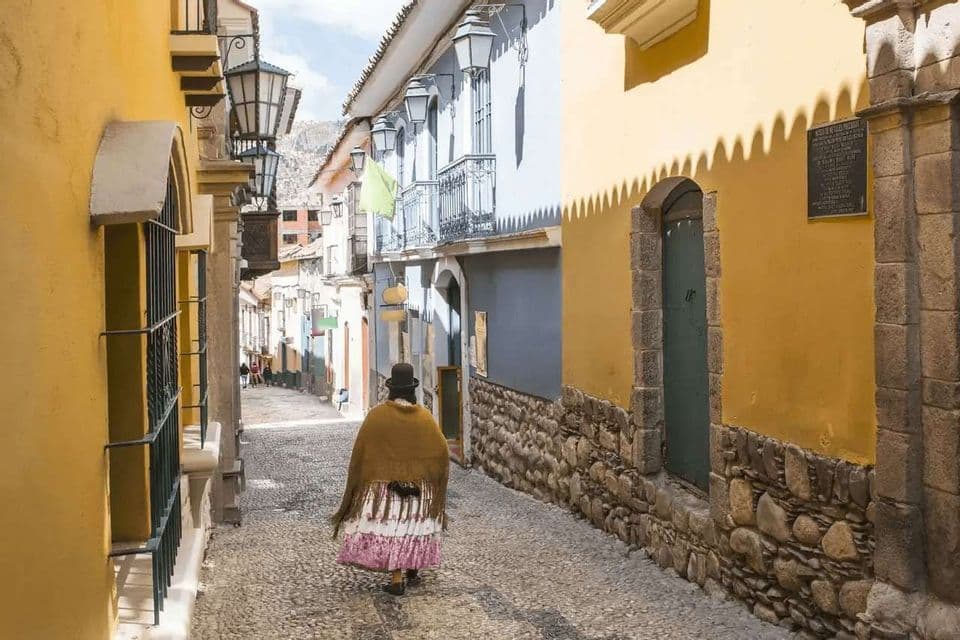 Una persona con bombín y mantón camina por un estrecho callejón empedrado entre edificios coloridos con balcones.