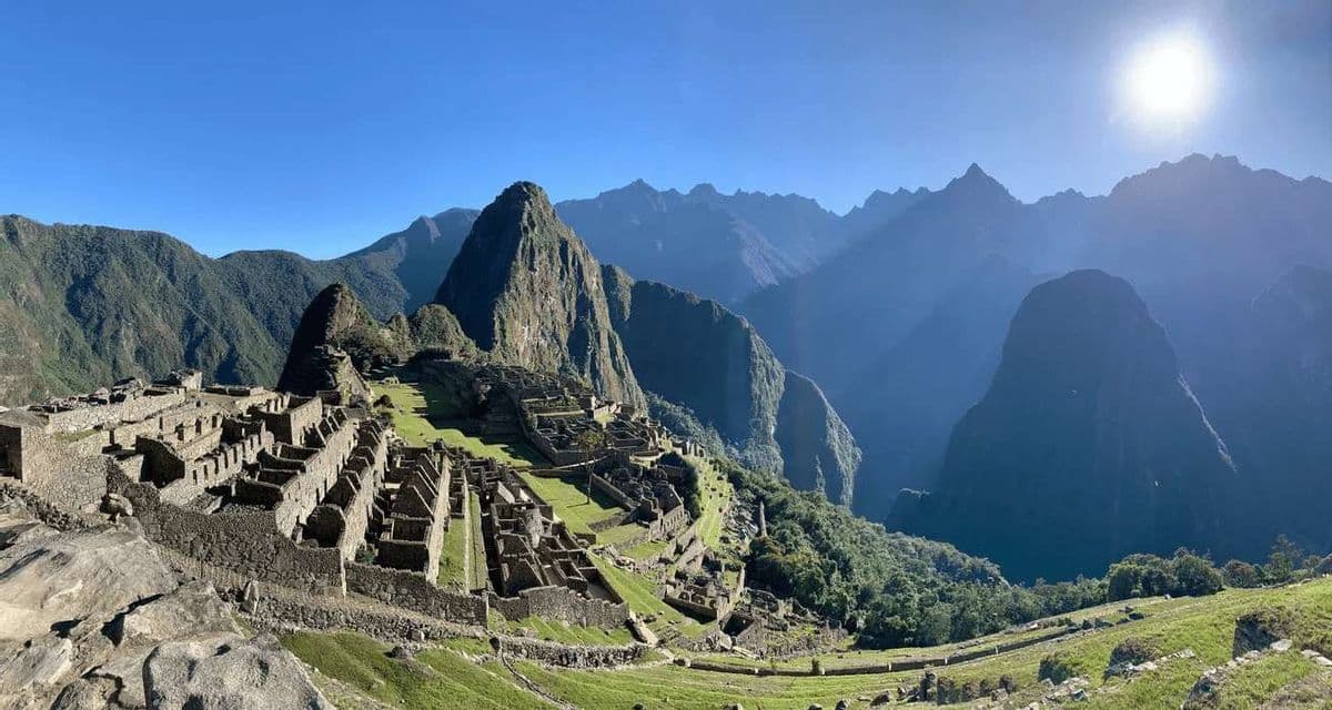 Una vista panoramica di antiche rovine in pietra su un fianco di montagna terrazzato, con vette ripide sullo sfondo sotto un sole splendente.