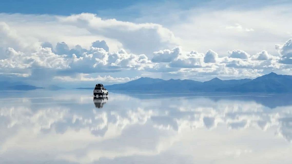 Un SUV blanco circula sobre un vasto salar reflectante, reflejando a la perfección el cielo nublado y las montañas distantes.