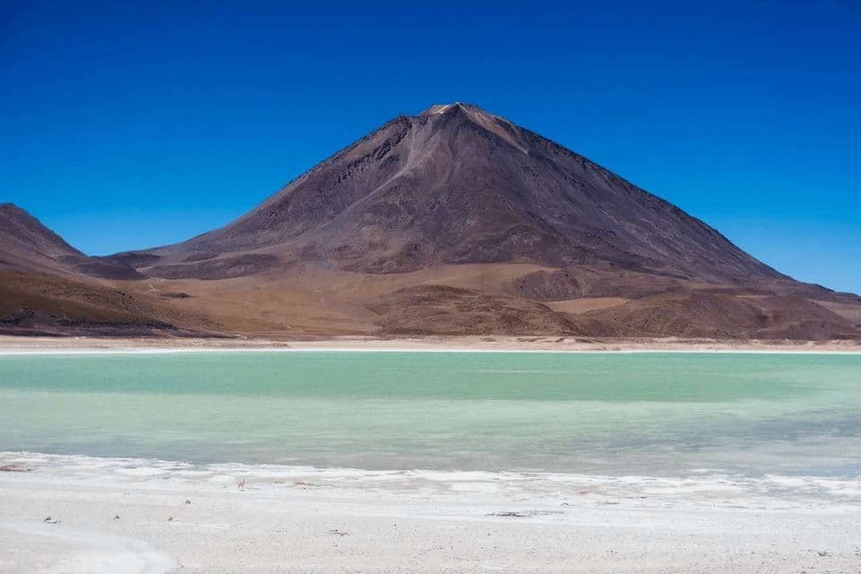 Una gran montaña marrón se alza detrás de un lago turquesa con una orilla blanca y salada bajo un cielo claro y azul.
