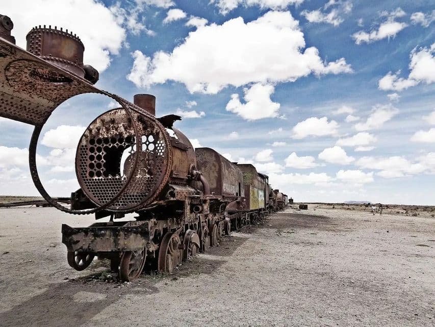Un long train à vapeur rouillé abandonné dans un paysage sec et plat sous un ciel bleu avec des nuages blancs.