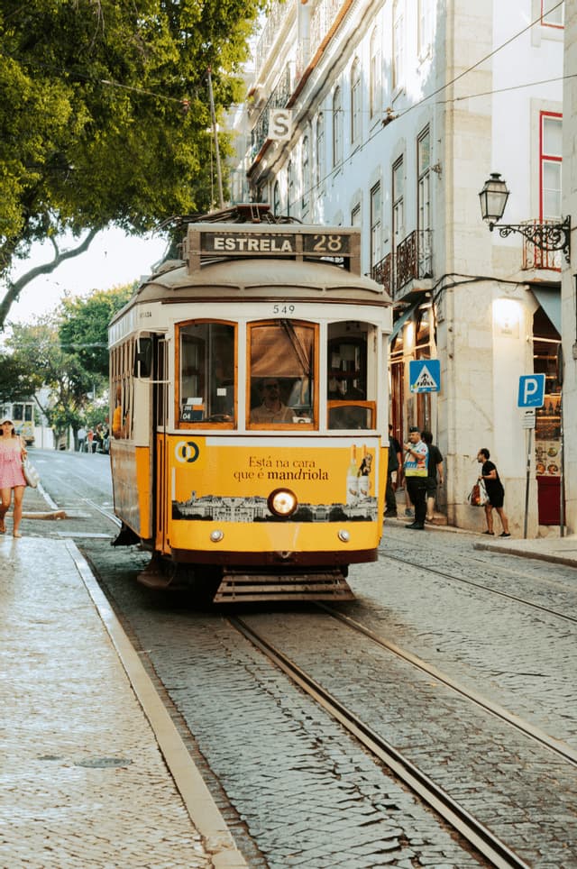 Un tram vintage giallo, numero 28, viaggia lungo i binari su una stretta strada di ciottoli costeggiata da edifici e alberi.