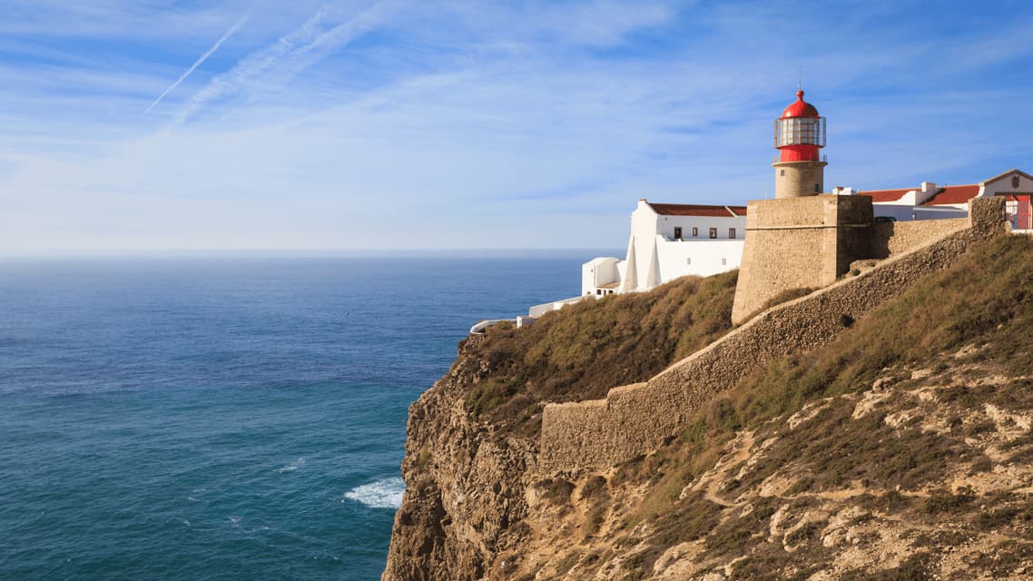 Un faro con tetto rosso e edifici bianchi sorge su una scogliera rocciosa affacciato sull'oceano in una giornata di sole.
