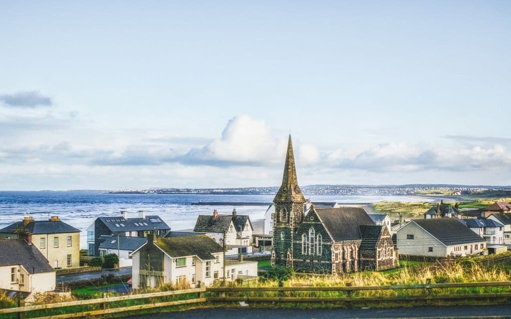 Una chiesa in pietra con un alto campanile domina un borgo costiero, con case situate in riva al mare, sotto un cielo parzialmente nuvoloso.