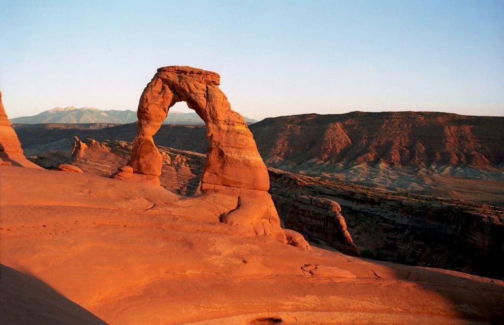 A large red sandstone arch stands in a rocky desert landscape, illuminated by the warm light of sunrise against a clear sky.