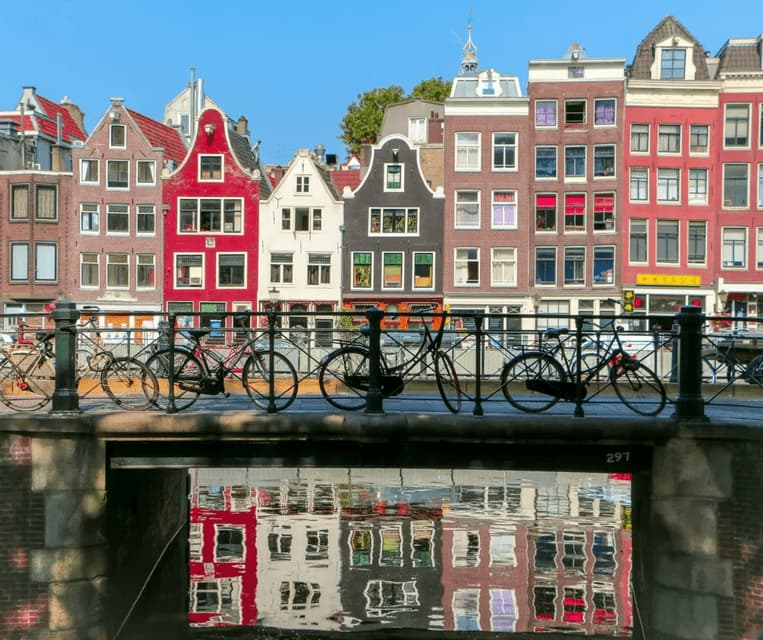 Una hilera de casas coloridas y con hastiales reflejadas en un canal, con bicicletas aparcadas en un puente en primer plano.