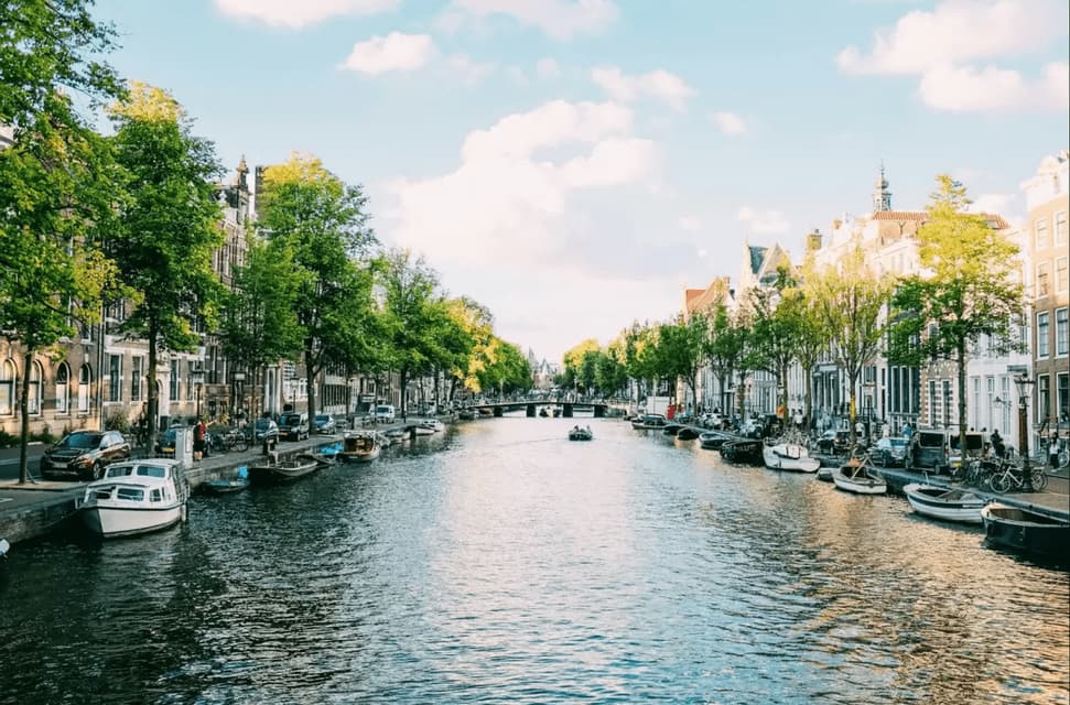 Una vista de un canal urbano con casas tradicionales, árboles verdes y barcos amarrados a sus orillas bajo un cielo azul con nubes blancas.