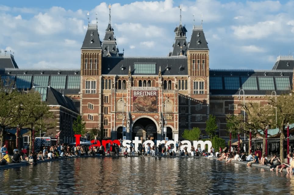 El letrero 'I amsterdam' se encuentra en un estanque reflectante frente al Rijksmuseum, con gente sentada a lo largo del borde.