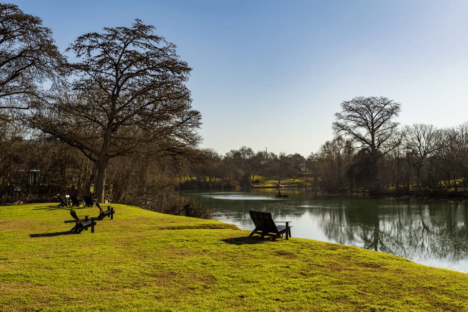 Empty black chairs on a grassy riverbank face a calm river with a person kayaking under a clear blue sky.