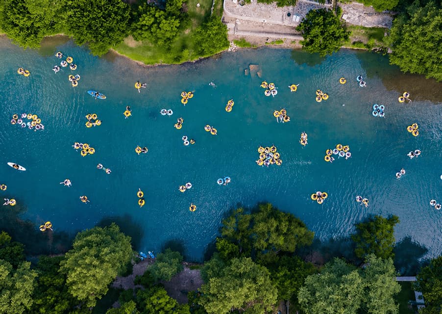 An aerial top-down view of a WeRoad group trip floating on inflatable rings down a blue river surrounded by green trees.