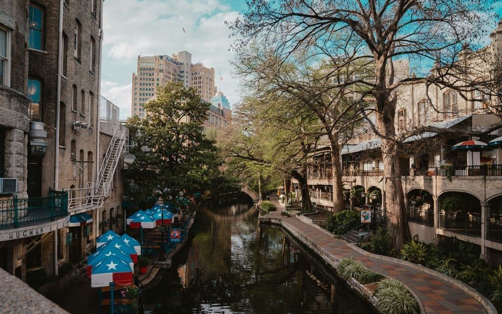 A city canal flows between brick buildings and a tree-lined walkway with restaurant patios and star-patterned umbrellas.