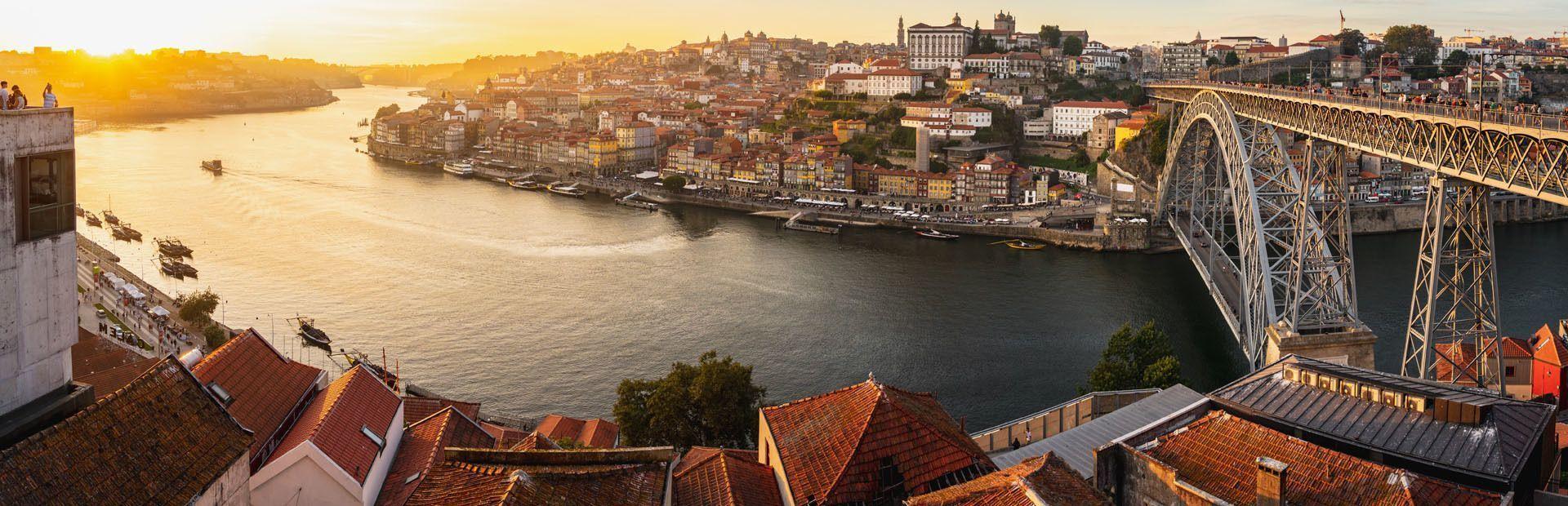 Panoramablick auf eine Flussstadt mit großer Metallbrücke und farbenfrohen Gebäuden im goldenen Licht des Sonnenuntergangs.