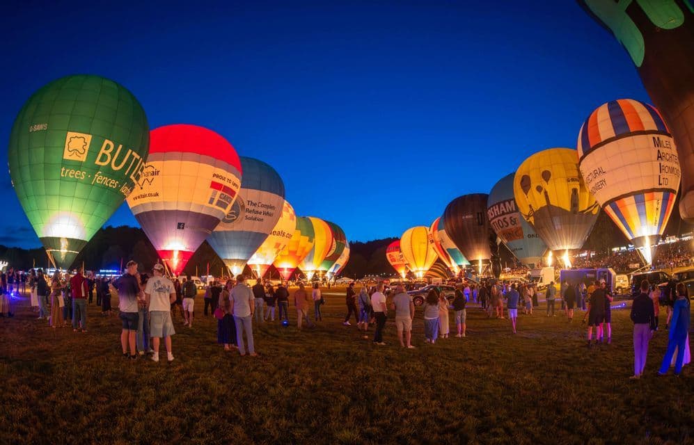 Mongolfiere colorate sono illuminate in fila su un campo erboso di notte, mentre una folla osserva.