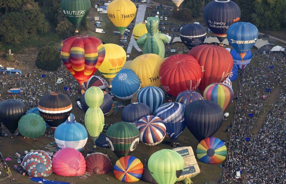 Uno scatto aereo di un grande campo ricolmo di decine di mongolfiere colorate durante un festival, circondato da una vasta folla di persone.