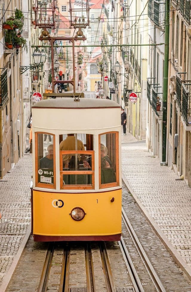 Un tram funicolare giallo con passeggeri a bordo risale una ripida e stretta strada acciottolata fiancheggiata da edifici.