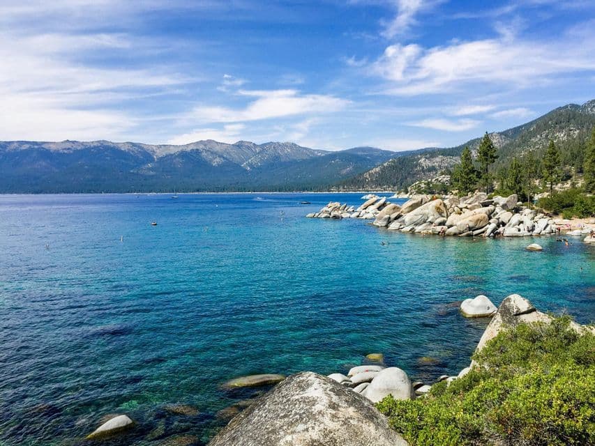 A clear, turquoise lake meets a rocky shoreline with large boulders, with forested mountains in the distance under a blue sky.
