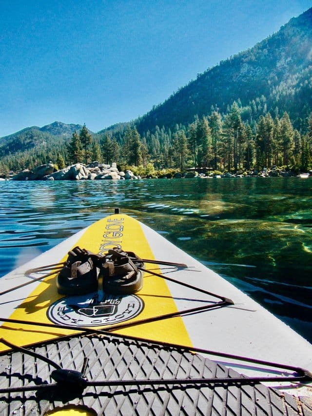 A first-person view from a yellow paddleboard with sandals on it, gliding across clear water toward a pine-covered shore.