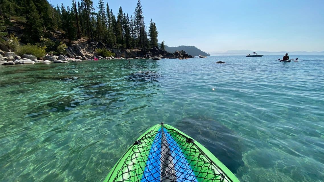 The bow of a green kayak floats on clear, turquoise water near a rocky shore with pine trees under a clear sky.