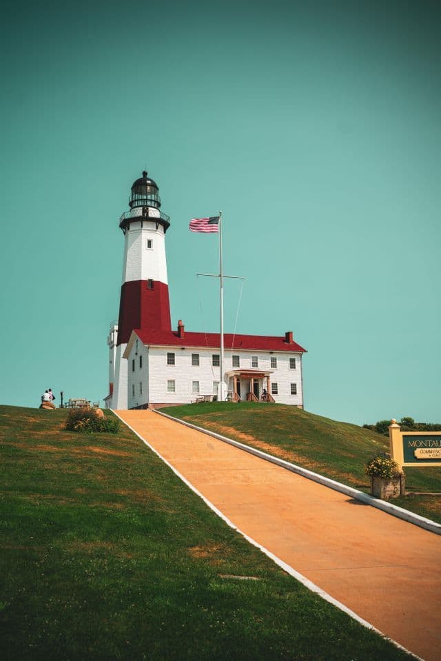 A red and white lighthouse with an American flag stands on a grassy hill under a teal sky, with a paved path leading towards it.