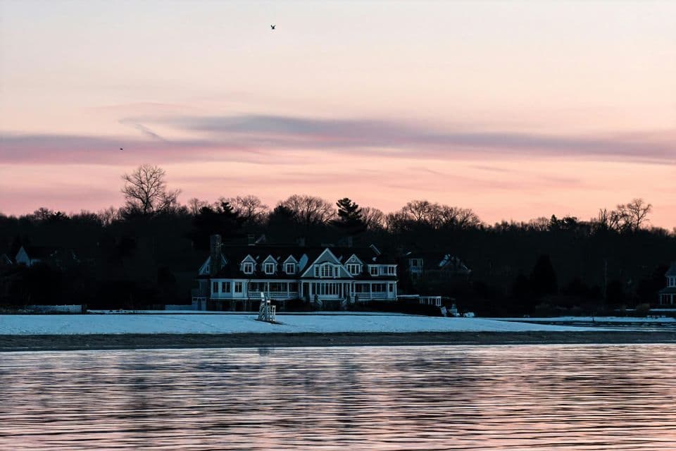 A large white house on a snowy shore viewed from across the water under a pink and purple sunset sky.