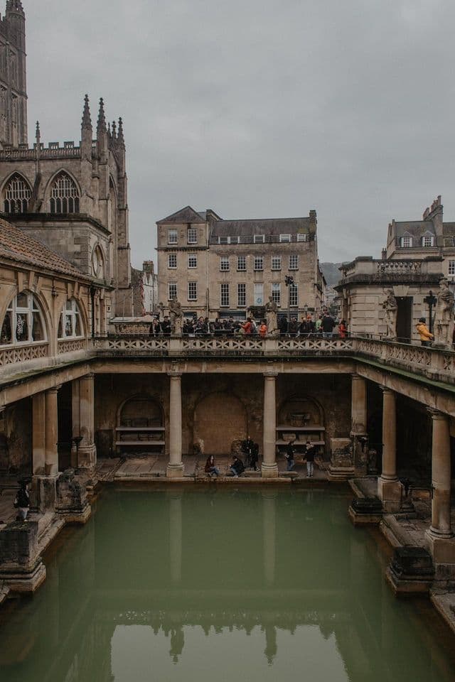 People on a terrace overlooking an ancient, stone-pillared bath with green water, with historic buildings under an overcast sky.