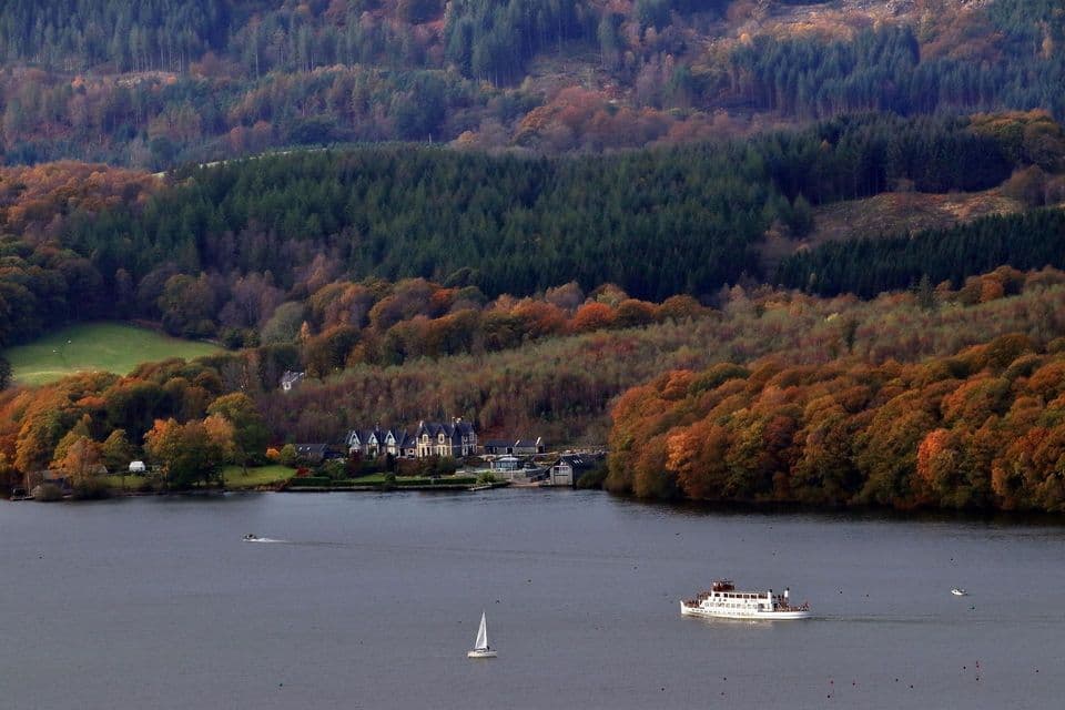 A ferry and other boats sail on a lake in front of a shore with houses and hills covered in autumn forests.
