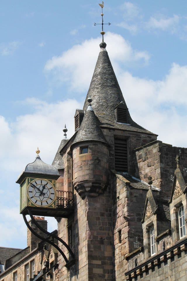A stone tower with a turreted, slate-tiled roof and a large, projecting clock, viewed against a cloudy blue sky.