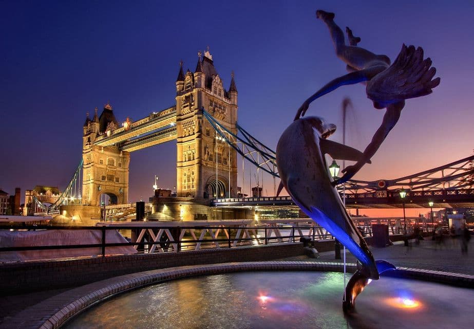 A dolphin fountain is illuminated in the foreground with a historic bridge lit up against a twilight sky in the background.
