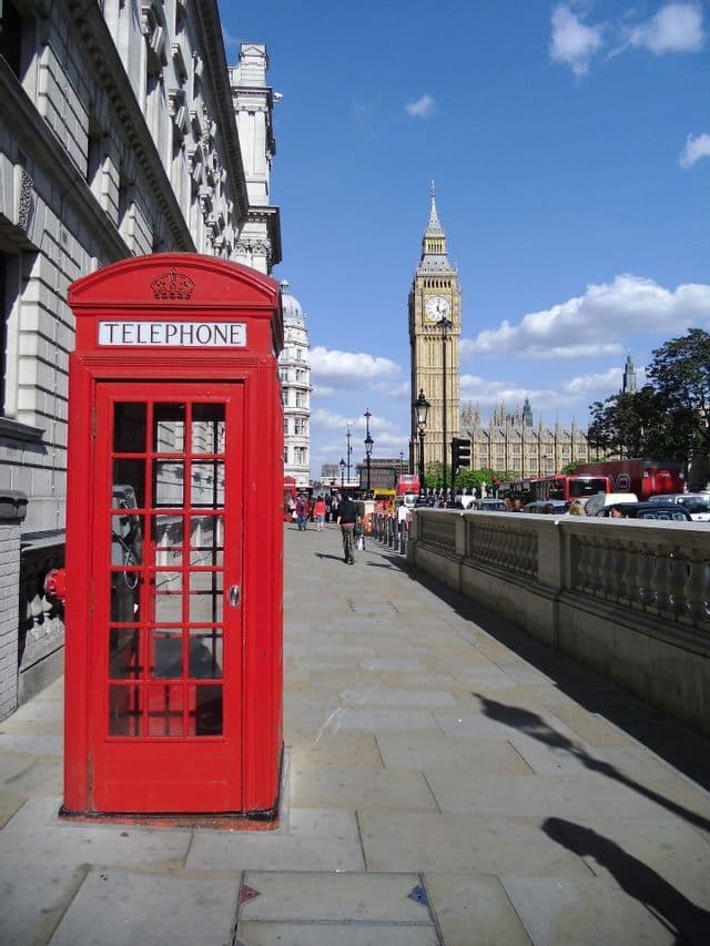 A classic red telephone booth on a city sidewalk with a large clock tower in the background under a blue sky.