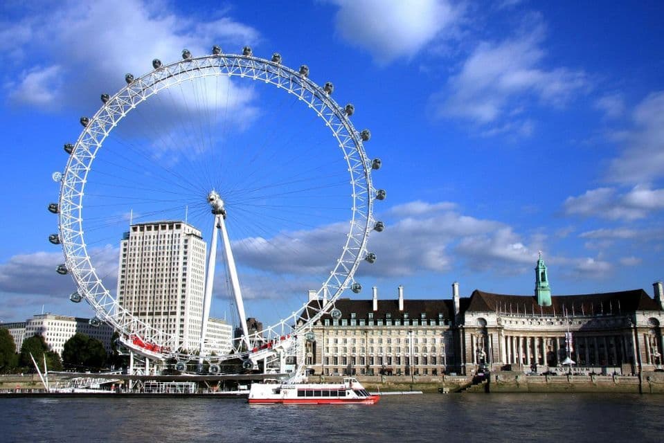 A large white Ferris wheel stands on a riverbank next to city buildings, with a boat in the water under a blue sky.