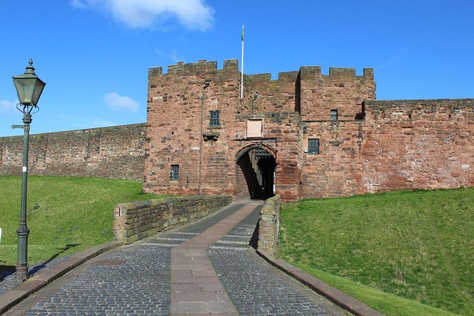 A cobblestone bridge leads to the arched entrance of a red brick castle gatehouse under a blue sky.