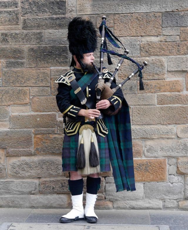 A man in traditional Scottish dress, including a tartan kilt and a bearskin hat, plays the bagpipes against a stone wall.