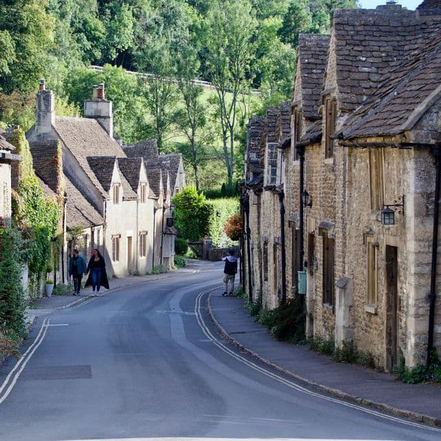 A winding road runs through a village of traditional stone cottages, with people walking on the sidewalks and a green hill in the background.