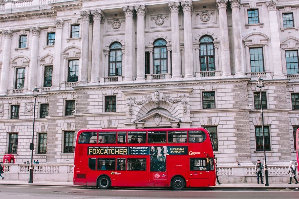 A red double-decker bus is parked on a street in front of a large, white classical building with columns.