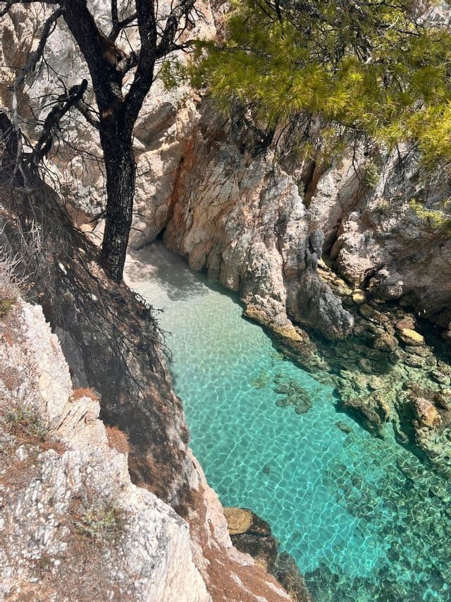 Vista dall'alto di una caletta appartata con piccola spiaggia sabbiosa e acqua cristallina turchese, incastonata tra scogliere rocciose e alberi.