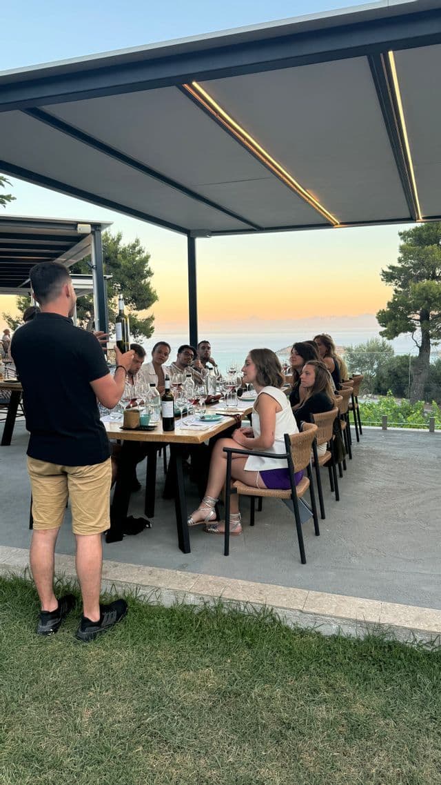 Un uomo presenta una bottiglia di vino a un gruppo WeRoad durante una degustazione su una terrazza all'aperto con vista mare al tramonto.