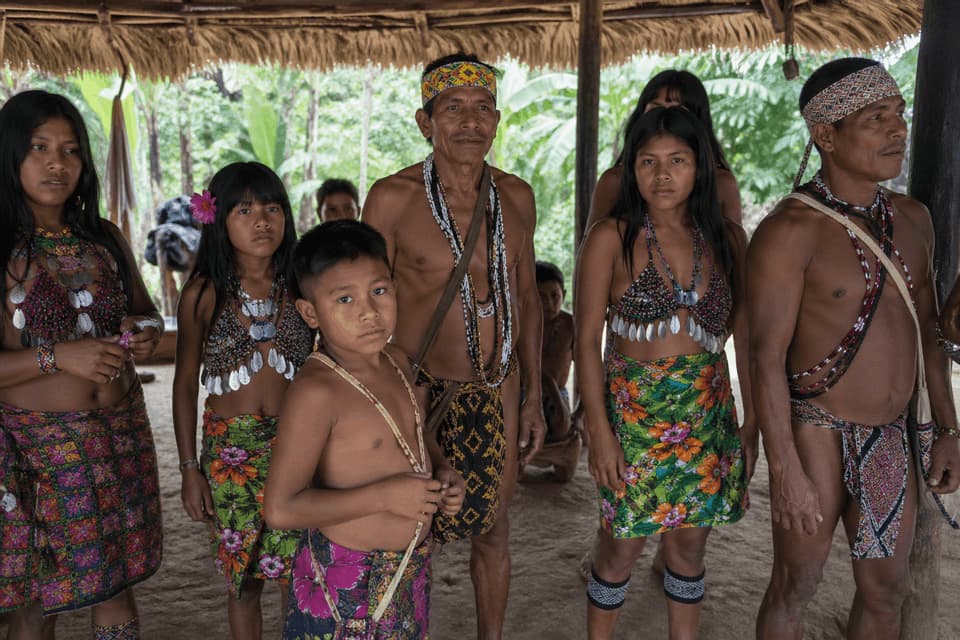 Un gruppo di indigeni, compresi adulti e bambini, sono riuniti in abiti tradizionali sotto una tettoia di paglia.