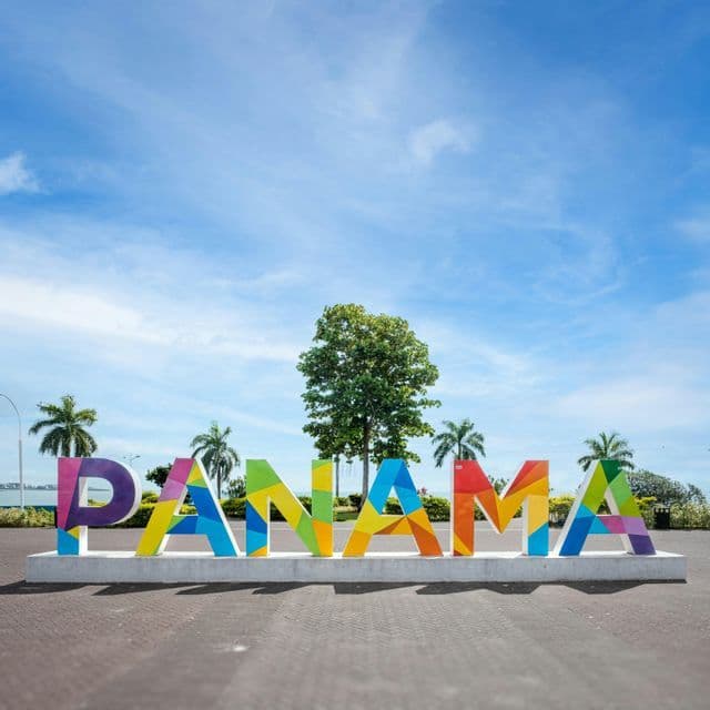 Un grande cartello colorato con la scritta 'PANAMA' si erge in una piazza sul lungomare, tra palme e sotto un cielo azzurro.
