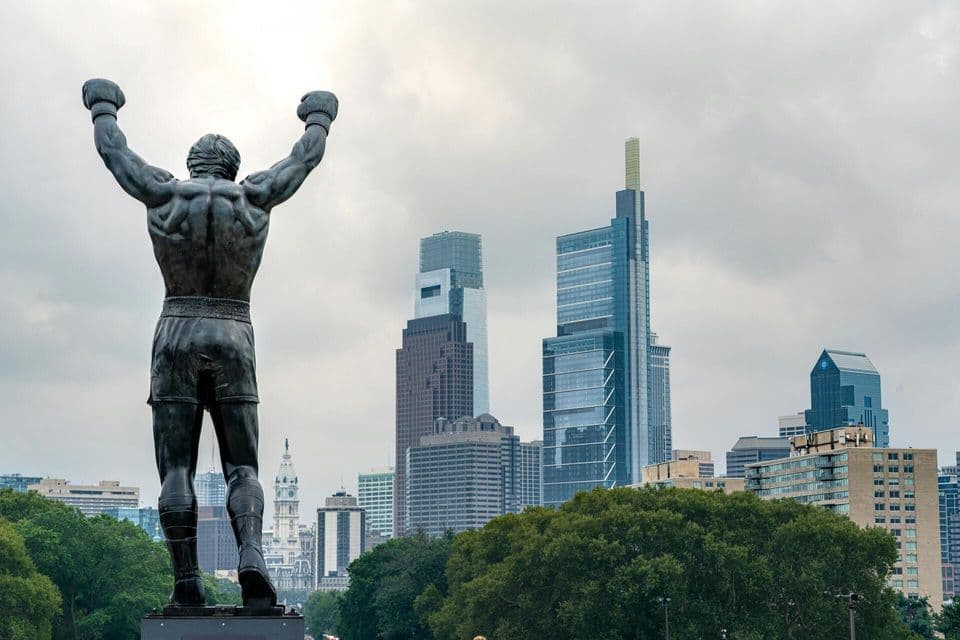 The back of a bronze statue of a boxer with its arms raised in a victory pose, overlooking a city skyline with skyscrapers.