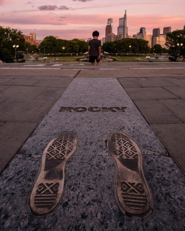 A low-angle view of bronze footprints and the word 'ROCKY' on a pavement, with a boy looking at a city skyline at sunset.