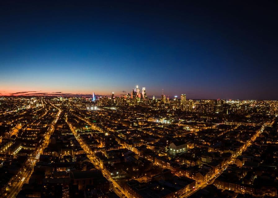 An aerial view of a sprawling city at dusk, with illuminated streets leading to a glowing skyline of skyscrapers.