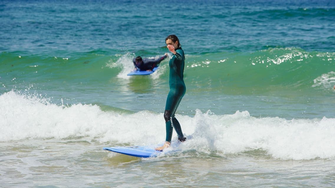 Una donna in muta verde sta su una tavola da surf blu, cavalcando una piccola onda durante un viaggio di gruppo WeRoad.