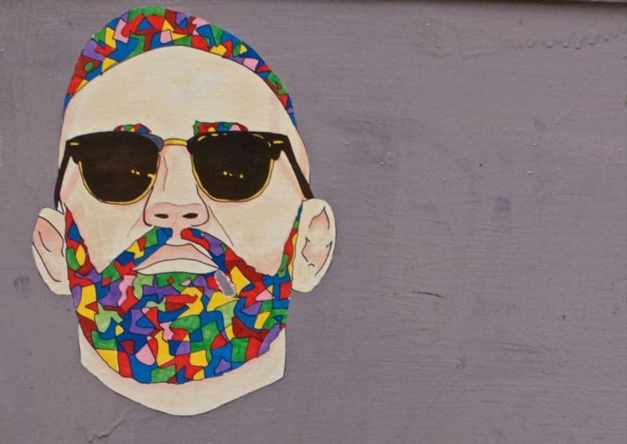 An illustration of a man's face with sunglasses, where his hair and beard are a colorful mosaic pattern, on a gray wall.