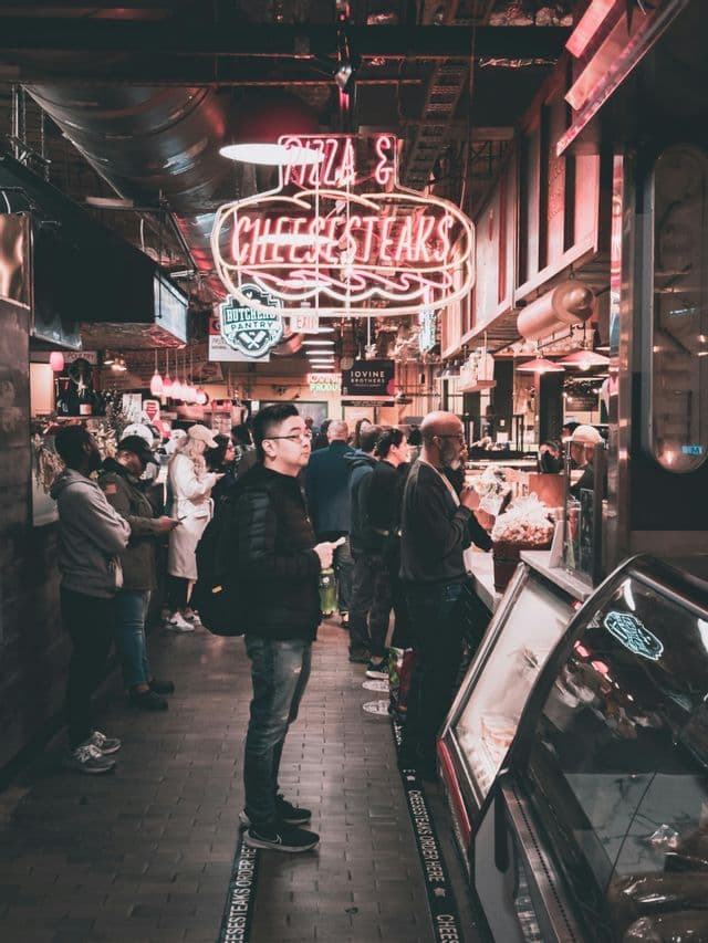 People stand in line at an indoor food market under a glowing red 'Pizza & Cheesesteaks' neon sign.