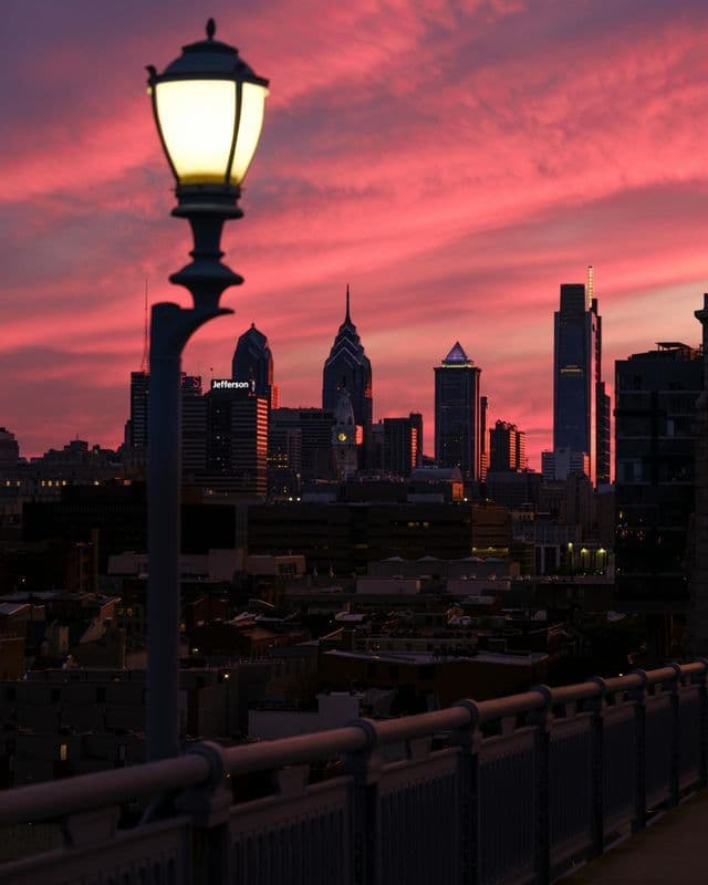 A lit street lamp in the foreground overlooks a city skyline silhouetted against a vibrant pink sunset.