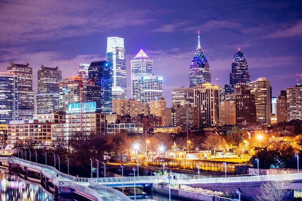 A city skyline with illuminated skyscrapers at twilight, with a modern pedestrian bridge crossing a river in the foreground.