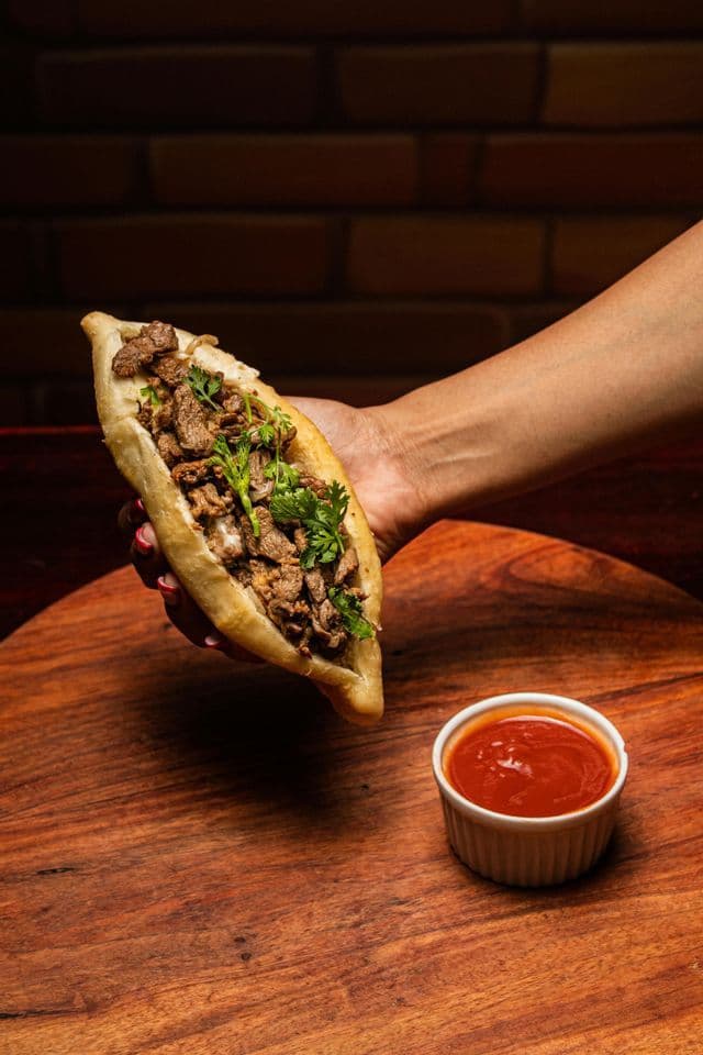 A hand holds a boat-shaped sandwich filled with meat and cilantro over a wooden board with a small bowl of red sauce.