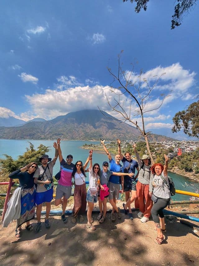 Un grupo de WeRoad posa con las manos en alto en un mirador, con vistas a un lago y una gran montaña bajo un cielo azul.